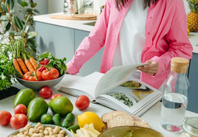 A person browsing a cookbook surrounded by fresh vegetables in a bright kitchen setting.